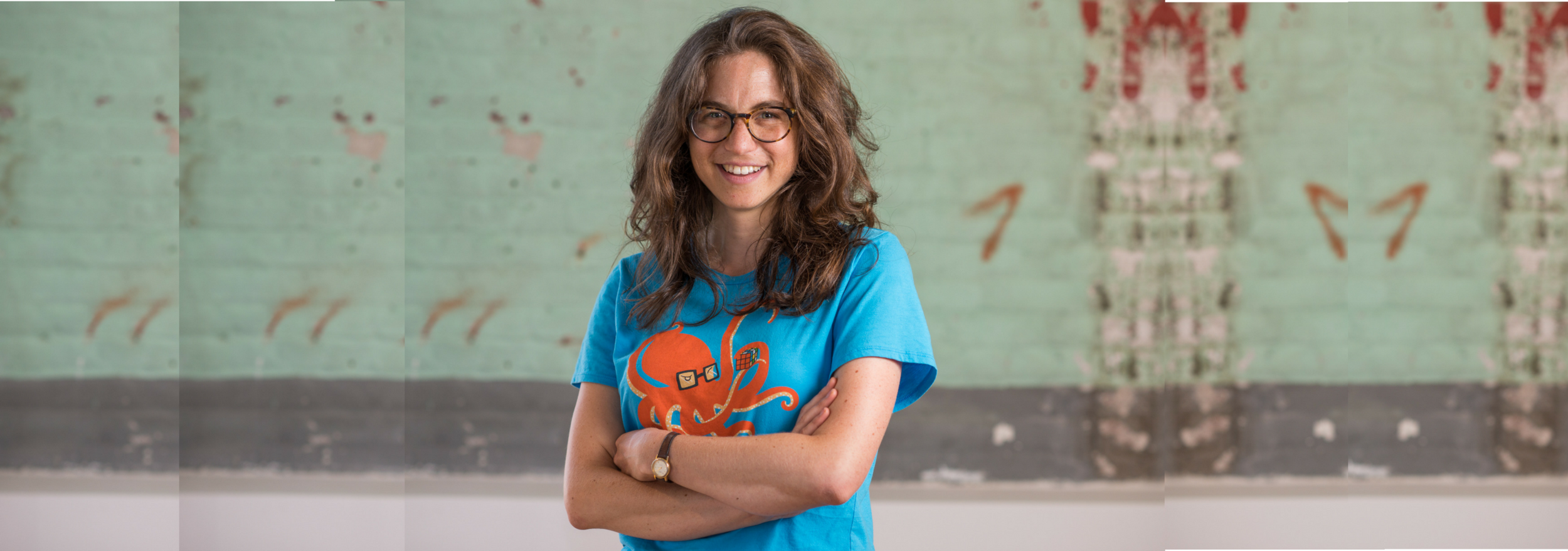 (Descrição da imagem: A reportér Wendy Zuckerman posa de braços cruzados em frente a um muro de tijolos azul claro. Ela está sorrindo e usando uma camisa com um desenho de um polvo de óculos)