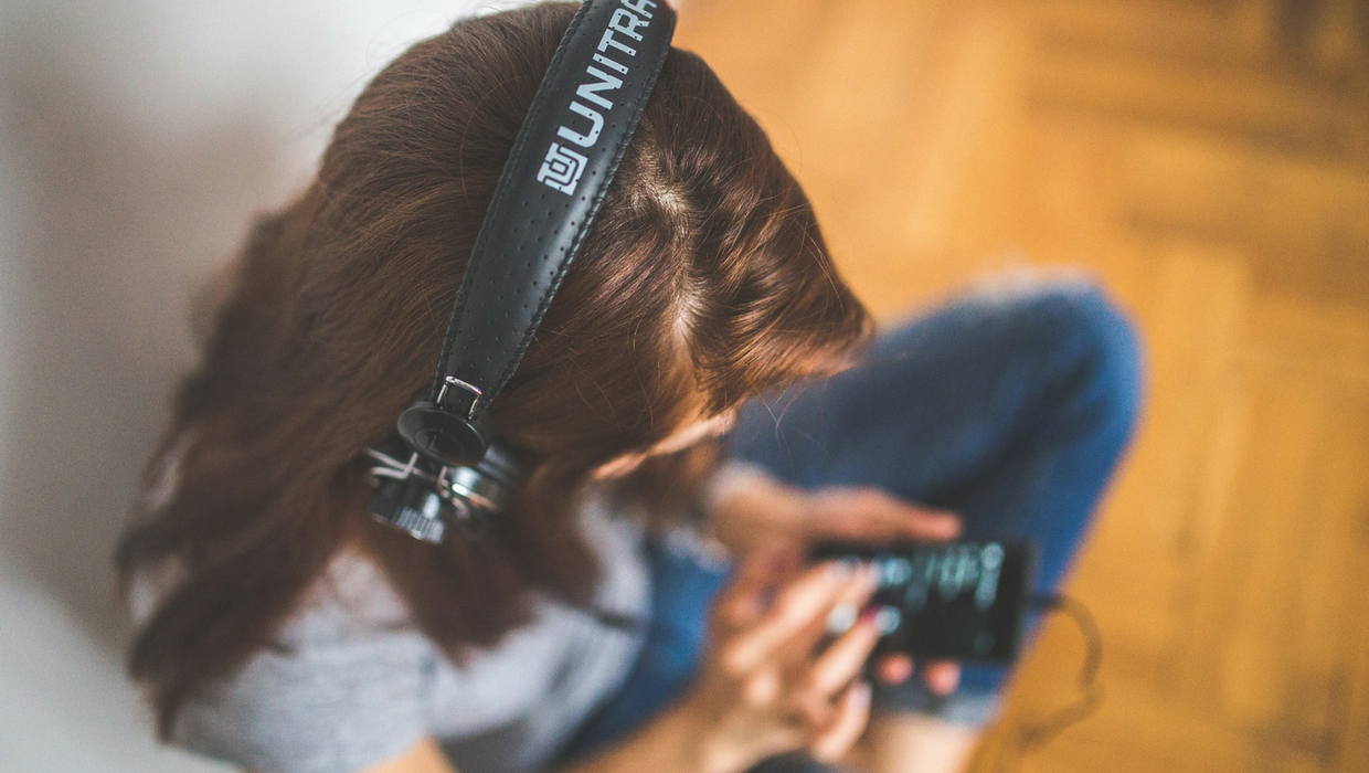 (Descrição da imagem: Mulher seleciona um conteúdo no celular. Ela está usando fones de ouvido ligados ao celular. A mulher está sentada no chão de madeira da sua casa)